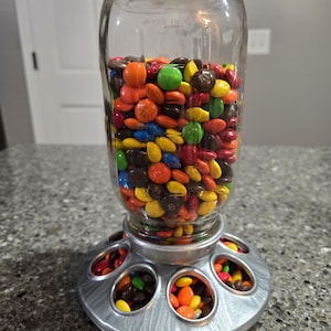 May include: A clear glass jar filled with colorful M&M's candies sits on a metal chicken feeder with multiple compartments. The feeder has a silver finish and is filled with M&M's.