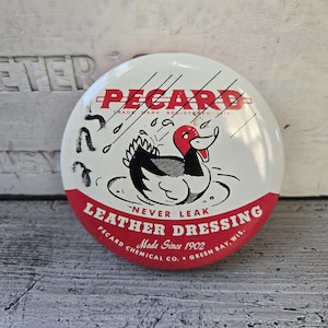 May include: A round, vintage PECARD Leather Dressing button with a white background and red trim. The button features a cartoon duck in a pond, with the text "NEVER LEAK" and "Made Since 1902".