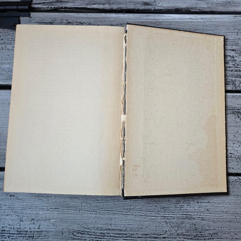 May include: An open book with aged, off-white pages. The binding is visible, showing the stitching. The book is lying on a weathered, gray wooden surface, suggesting a vintage or antique item. The pages are blank, ready for writing or display.
