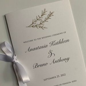 May include: White wedding program with gold lettering announcing the wedding ceremony of Anastasia Kathleen and Bruno Anthony. The ceremony will take place on September 25, 2022 at the Holy Monastery of Panagia, Kera, Crete, Greece.
