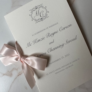 May include: A white wedding invitation with a black monogram of "MC" in a decorative frame. The invitation announces the marriage of Mc Kenzie Rayne Carson and terios Channing Stroud on November 25, 2023 at St. Paul's Greek Orthodox Church in Savannah, Georgia.