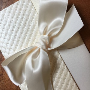 May include: A white card with a white satin ribbon tied in a bow. The card has a textured pattern.