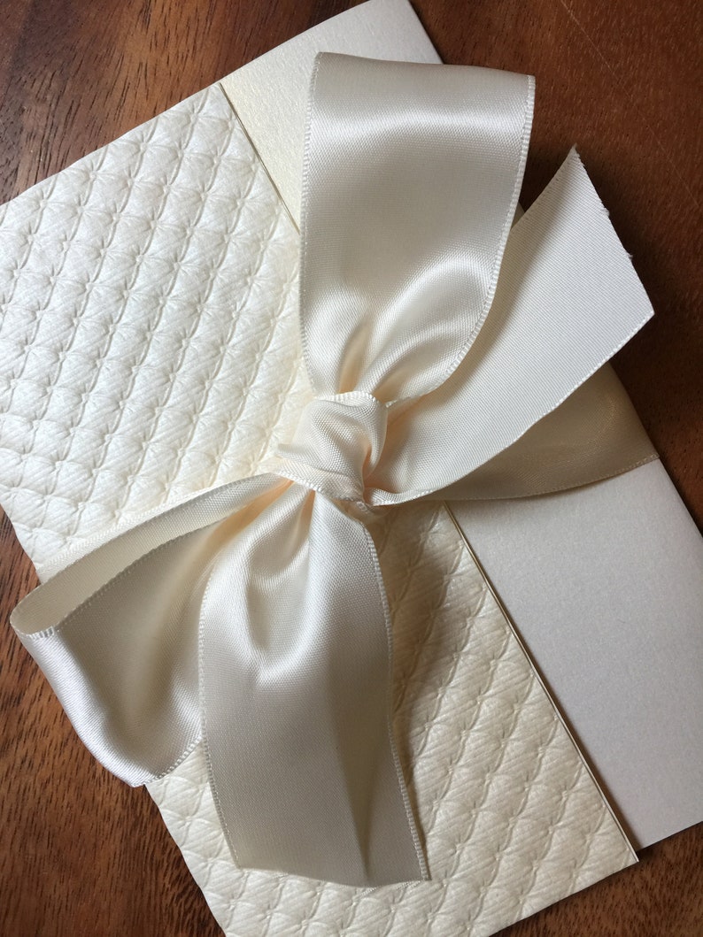 May include: A white card with a white satin ribbon tied in a bow. The card has a textured pattern.