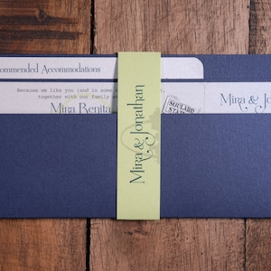 May include: Navy blue wedding invitation suite with a light green belly band. The invitation includes text that reads "Mira & Jonathan" and "Mira & Jon Wedding 2016". The suite is on a wooden surface.