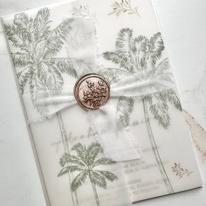 May include: A translucent vellum invitation card featuring green palm tree illustrations, tied with a white frayed-edge ribbon and secured by a circular metallic rose gold wax seal embossed with a floral design, displaying printed text details.