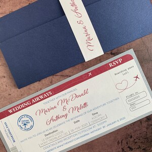 May include: A navy blue wedding invitation suite featuring a boarding pass design. The invitation includes the names "Marina & Anthony", date, time, and location details. A matching belly band with the names "Marina & Anthony" wraps the invitation.