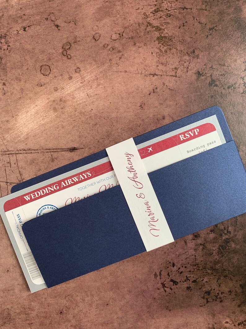 May include: Navy blue wedding invitation designed to resemble an airline boarding pass. The invitation includes the text "WEDDING AIRWAYS" and "Marina & Anthony" in burgundy script. A white band wraps around the invitation, also bearing the names.