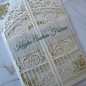 May include: Ivory wedding invitation with a laser-cut gate design and gold accents. The invitation is held together with a translucent band printed with the names "Kaylee Andrea Fabian" in green script. The overall design is elegant and ornate.