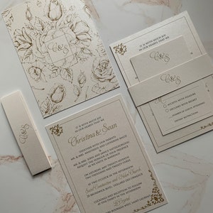 May include: Elegant wedding invitation suite in cream with gold floral accents. The suite includes a main invitation, RSVP card, enclosure card, and a belly band, all featuring the initials "C&S". The main invitation includes the names Christina & Sean.