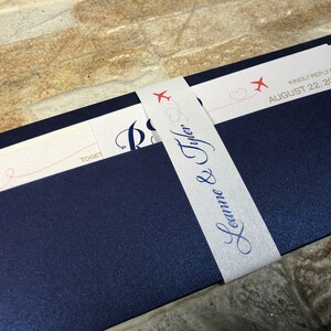 May include: Navy blue wedding invitation suite with a white belly band. The invitation features the names "Leanne & Tyler" in elegant script. The band is decorated with red airplane graphics and the text "Kindly reply by August 22, 2018."