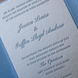 May include: A light blue wedding invitation with embossed text. The invitation includes the names Jessica Louise and Griffin Lloyd Andrew, along with the date, time, and location of the wedding ceremony.