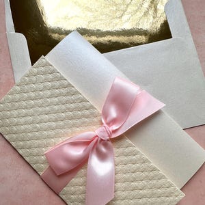 May include: A cream-colored envelope with a pink satin ribbon tied in a bow. The envelope has a textured pattern and is nestled inside a white envelope with a gold inner lining.