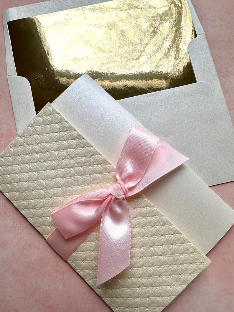 May include: A cream-colored envelope with a pink satin ribbon tied in a bow. The envelope has a textured pattern and is nestled inside a white envelope with a gold inner lining.