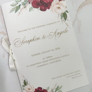 May include: A white wedding ceremony programme booklet featuring burgundy and cream floral watercolour illustrations at the top and bottom. The text reads: Welcome to the wedding ceremony of Seraphim & Angela, Sunday, August 16, 2026, at 3:00pm, St. Demetrios Greek Orthodox Church, Camarillo, California, and a quote from Matthew 19:6. A white ribbon is tied on the left side.