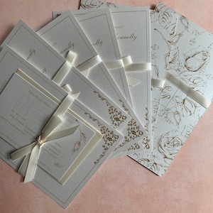 May include: Cream-colored wedding invitations with gold floral accents and the name "Connolly" in elegant script. Each invitation is tied with a cream ribbon. Some feature rose patterns, others ornate borders.