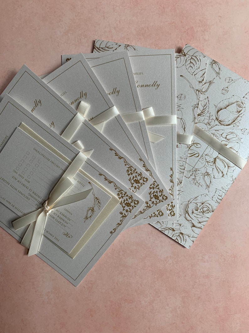 May include: Cream-colored wedding invitations with gold floral accents and the name "Connolly" in elegant script. Each invitation is tied with a cream ribbon. Some feature rose patterns, others ornate borders.