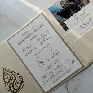 May include: A white and gold wedding invitation with Arabic calligraphy and a photo of a building. The invitation includes the names of the couple, the date and time of the wedding, and the location. The RSVP details are on a separate card.