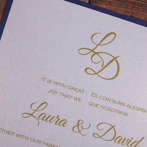 May include: A wedding invitation with a white background and navy blue border. The invitation features a gold monogram of the letters "L" and "D" in a script font. The text "It is with great joy that we" and "Es con suma alegr&iacute;a que nosotros" is printed in gold below the monogram. The names "Laura & David" are printed in gold below the text.