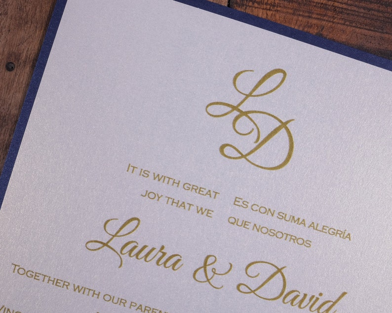 May include: A wedding invitation with a white background and navy blue border. The invitation features a gold monogram of the letters "L" and "D" in a script font. The text "It is with great joy that we" and "Es con suma alegr&iacute;a que nosotros" is printed in gold below the monogram. The names "Laura & David" are printed in gold below the text.