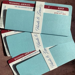 May include: Wedding invitation cards designed as boarding passes. The cards are light blue with a red and white top section that reads "Wedding Airways". Each card is wrapped with a white band printed with the names "Kaede & Lorne". RSVP details are also included.