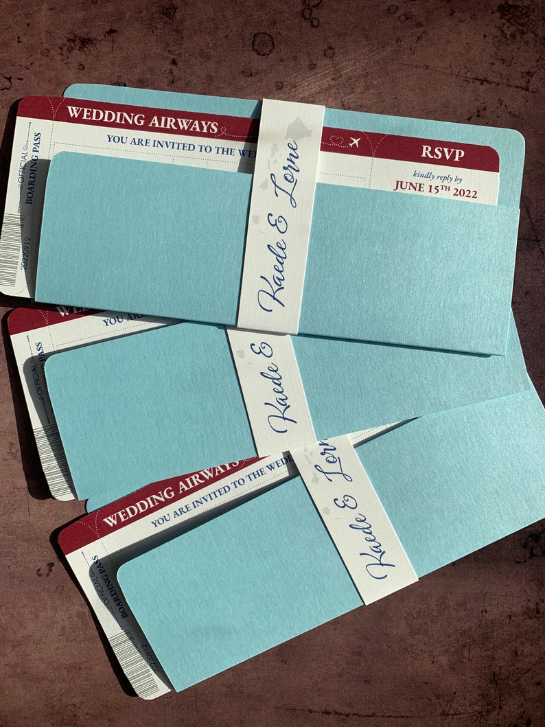 May include: Wedding invitation cards designed as boarding passes. The cards are light blue with a red and white top section that reads "Wedding Airways". Each card is wrapped with a white band printed with the names "Kaede & Lorne". RSVP details are also included.