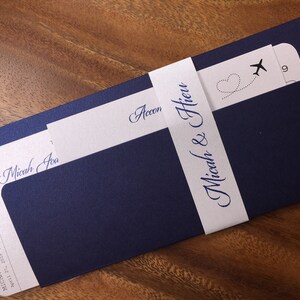 May include: Navy blue wedding invitation suite with a boarding pass design. The invitation features the names "Micah & Hieu" and the date April 27, 2019. A white band wraps around the invitation with the names in elegant script.