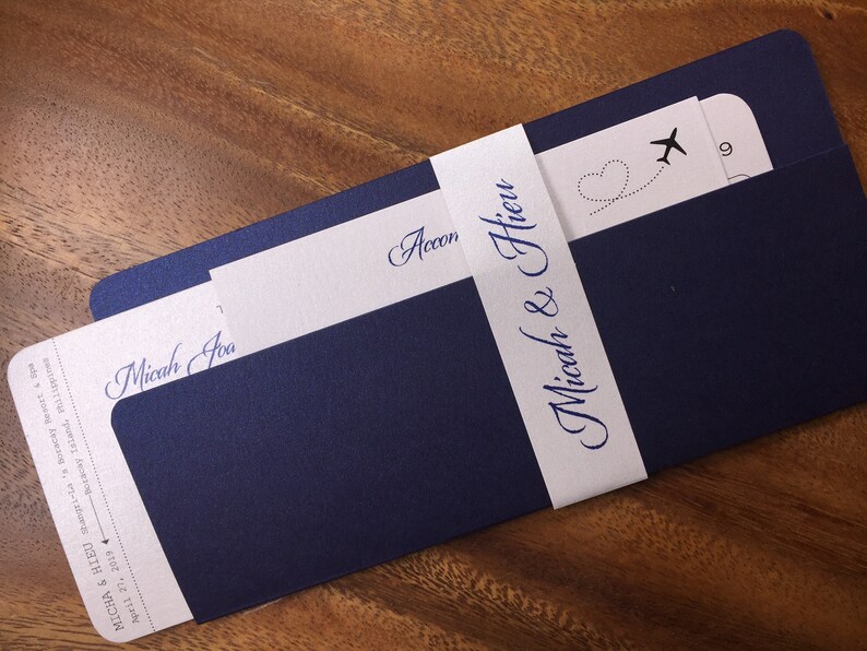 May include: Navy blue wedding invitation suite with a boarding pass design. The invitation features the names "Micah & Hieu" and the date April 27, 2019. A white band wraps around the invitation with the names in elegant script.