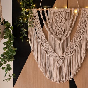 May include: A beige macrame wall hanging with a geometric pattern and fringe. The hanging is decorated with string lights and is displayed on a wooden wall.