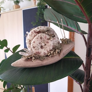Puede incluir: Un caracol de tela hecho a mano con un patrón floral y un acento de perla. El caracol está sentado en una hoja verde.
