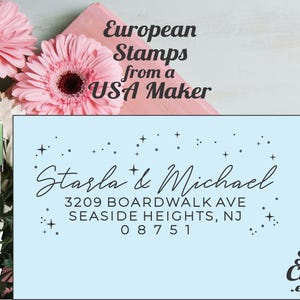 May include: A custom stamp with the names "Starla & Michael" and address details. The design includes a wooden stamp, a clear stamp, and the text "European Stamps from a USA Maker". The background features pink flowers.