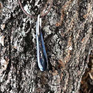 Puede incluir: Un collar con un colgante en forma de lágrima de color azul oscuro y dorado con diseño de mármol. El colgante está colgado de un cordón marrón.