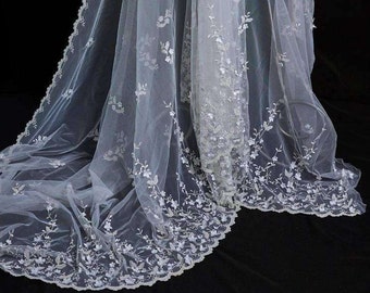May include: White sheer fabric with delicate floral embroidery and a scalloped edge. The fabric is draped over a dress form.