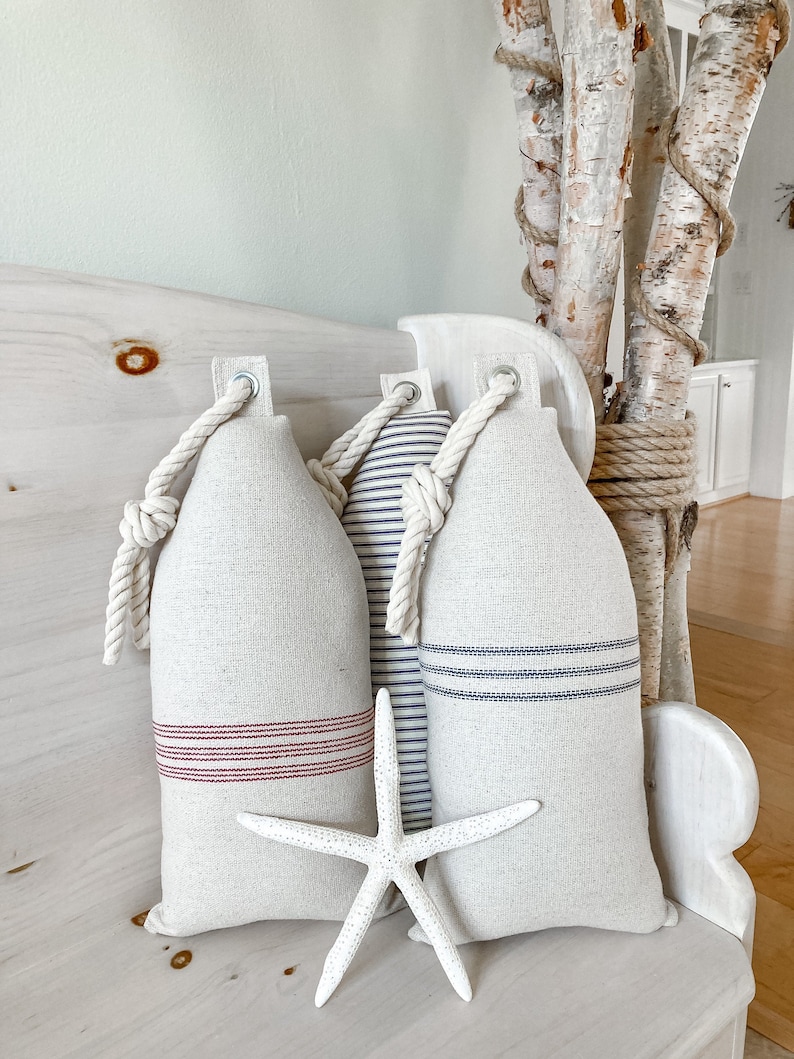 May include: Three decorative pillows with a nautical theme. The pillows are made of a natural linen fabric with red and blue stripes. The pillows are tied with rope and have a rope handle. A white starfish is in front of the pillows.