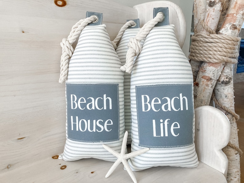 May include: Two white and gray striped doorstops with rope handles. The doorstop on the left has a gray patch with the text "Beach House" in white. The doorstop on the right has a gray patch with the text "Beach Life" in white.