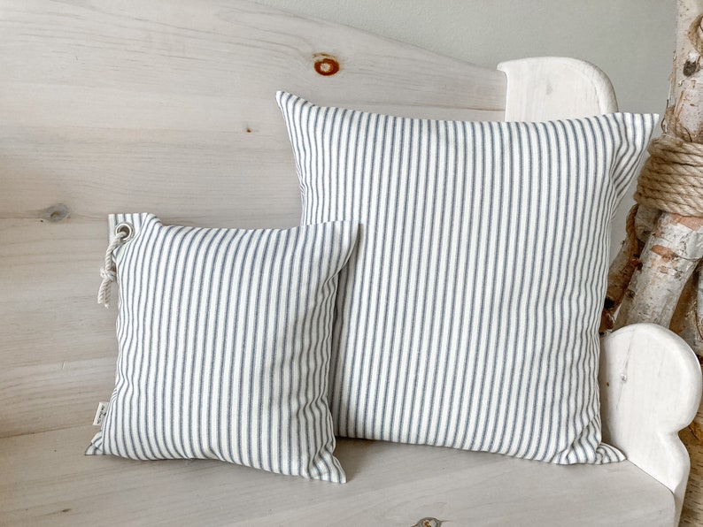 May include: Two striped throw pillows, one larger than the other, with a white background and thin blue stripes. The pillows are sitting on a white wooden bench.