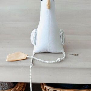May include: A white stuffed goose toy with a light yellow beak and feet. The toy has small black eyes and striped wings. A white rope is attached to the goose, with a light yellow tag at the end. The toy is sitting on a white wooden surface.
