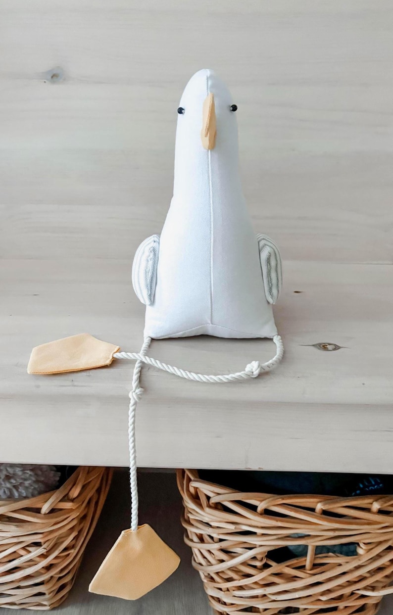 May include: A white stuffed goose toy with a light yellow beak and feet. The toy has small black eyes and striped wings. A white rope is attached to the goose, with a light yellow tag at the end. The toy is sitting on a white wooden surface.