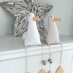 May include: Two plush seagull figurines with beige beaks and dangling legs. One is white, the other is light gray. Each has a rope leg with a bell and a tan tag. A large driftwood star is in the background.