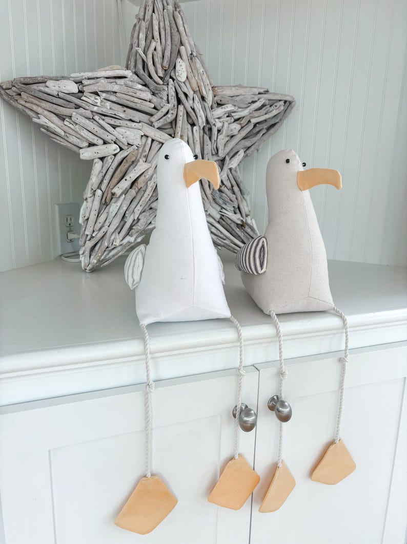 May include: Two plush seagull figurines with beige beaks and dangling legs. One is white, the other is light gray. Each has a rope leg with a bell and a tan tag. A large driftwood star is in the background.