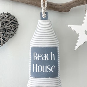 May include: A gray and white striped doorstop with a gray fabric patch that says "Beach House" in white text. The doorstop is hanging from a rope and a wooden knob.
