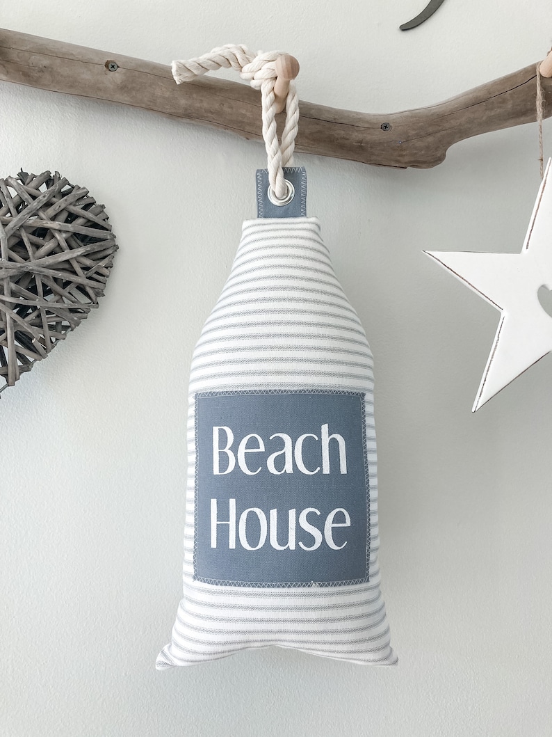 May include: A gray and white striped doorstop with a gray fabric patch that says "Beach House" in white text. The doorstop is hanging from a rope and a wooden knob.