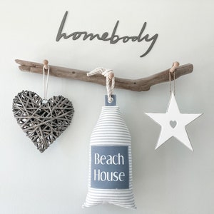 May include: A driftwood branch with three hanging decorations: a woven heart, a white star with a heart cutout, and a striped fabric pouch with the text "Beach House". The word "homebody" is written in black script above the branch.