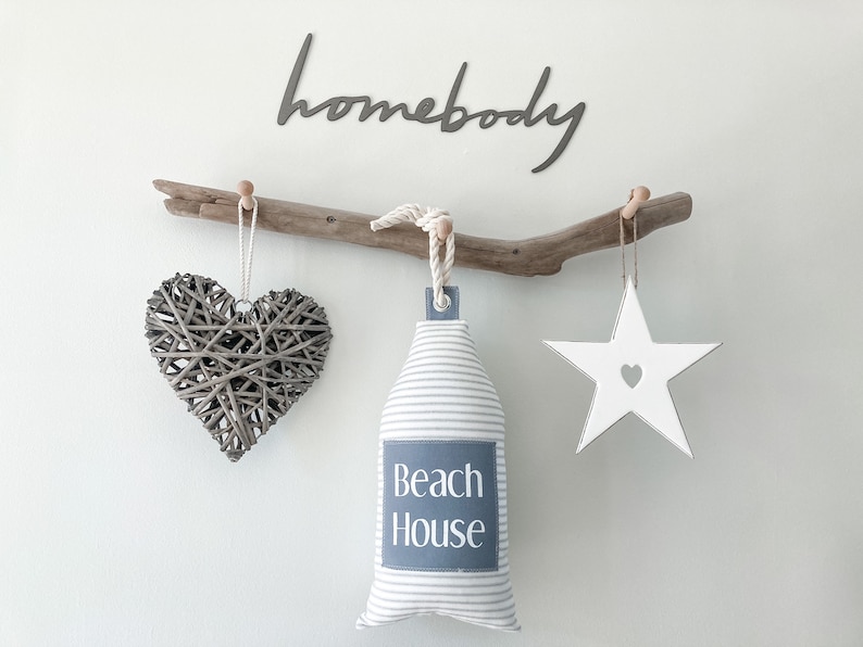 May include: A driftwood branch with three hanging decorations: a woven heart, a white star with a heart cutout, and a striped fabric pouch with the text "Beach House". The word "homebody" is written in black script above the branch.