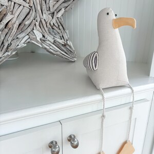 May include: A stuffed seagull with a beige body, a yellow beak, and black and white striped wing detail. The seagull has dangling legs with tan feet. A driftwood star is in the background.