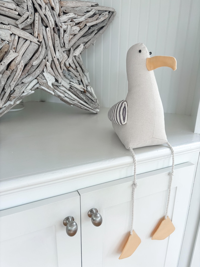 May include: A stuffed seagull with a beige body, a yellow beak, and black and white striped wing detail. The seagull has dangling legs with tan feet. A driftwood star is in the background.