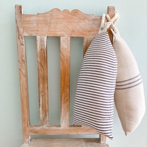 May include: Two decorative pillows with striped patterns hang from a wooden chair. The chair is light brown with a distressed finish. The pillows are made of a soft, natural fabric. One pillow has white and black stripes, and the other has a beige and brown striped pattern.