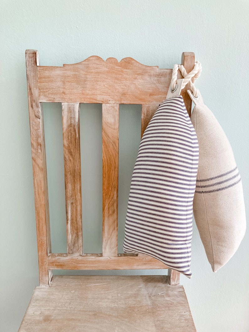 May include: Two decorative pillows with striped patterns hang from a wooden chair. The chair is light brown with a distressed finish. The pillows are made of a soft, natural fabric. One pillow has white and black stripes, and the other has a beige and brown striped pattern.