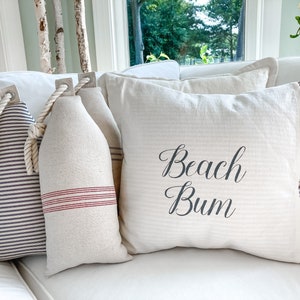 May include: Three decorative pillows on a white couch. One pillow is white with red stripes and has a rope handle. Another pillow is white with black stripes and has a rope handle. The third pillow is white with the text "Beach Bum" in black.