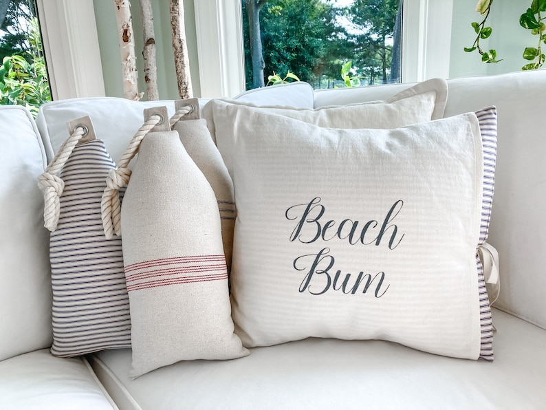 May include: Three decorative pillows on a white couch. One pillow is white with red stripes and has a rope handle. Another pillow is white with black stripes and has a rope handle. The third pillow is white with the text "Beach Bum" in black.