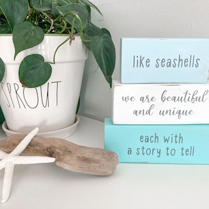 Puede incluir: Tres bloques de madera apilados uno encima del otro. El bloque superior es azul claro y dice "como conchas marinas". El bloque del medio es blanco y dice "somos hermosos y únicos". El bloque inferior es turquesa y dice "cada uno con una historia que contar".